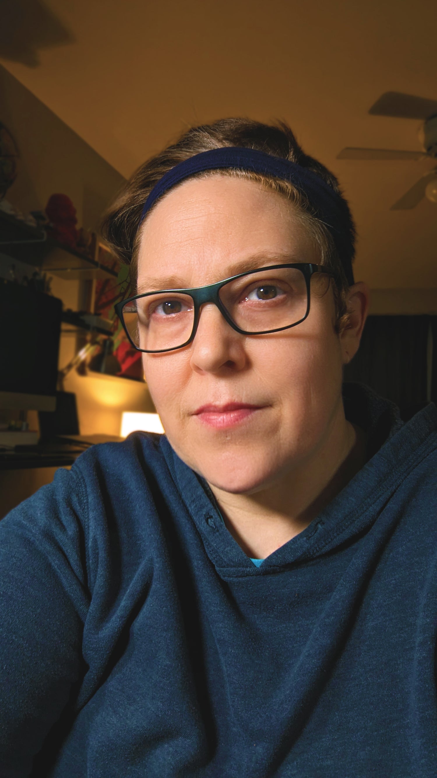 This is a portrait photo showing the face and shoulders of artist Katy V. Meehan. She is wearing a dark blue hooded sweatshirt, glasses, and a black headband over show auburn hair.